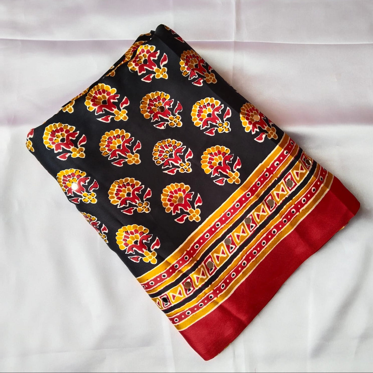 A folded black fabric saree or cloth featuring a repeating red and yellow floral block print and a decorative red, yellow, and black border, displayed on a white fabric background.