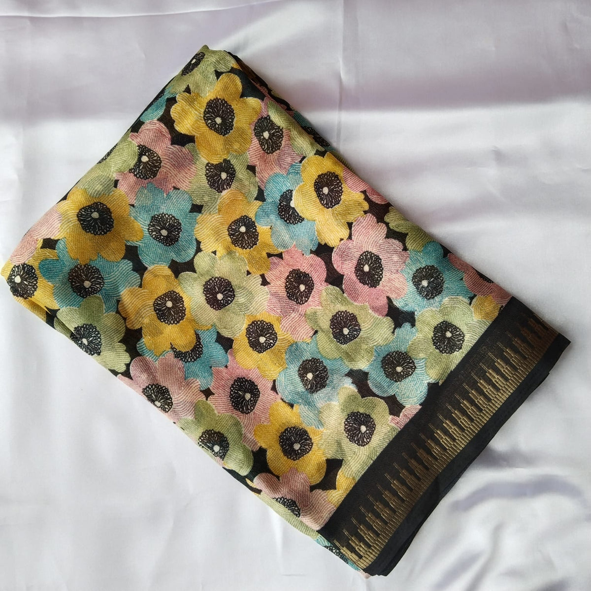 A folded multicolor floral printed fabric, likely a saree, laid flat on a white fabric background. The pattern features large flowers in yellow, pink, and light blue with black centers on a black base, finished with a black and gold patterned border.